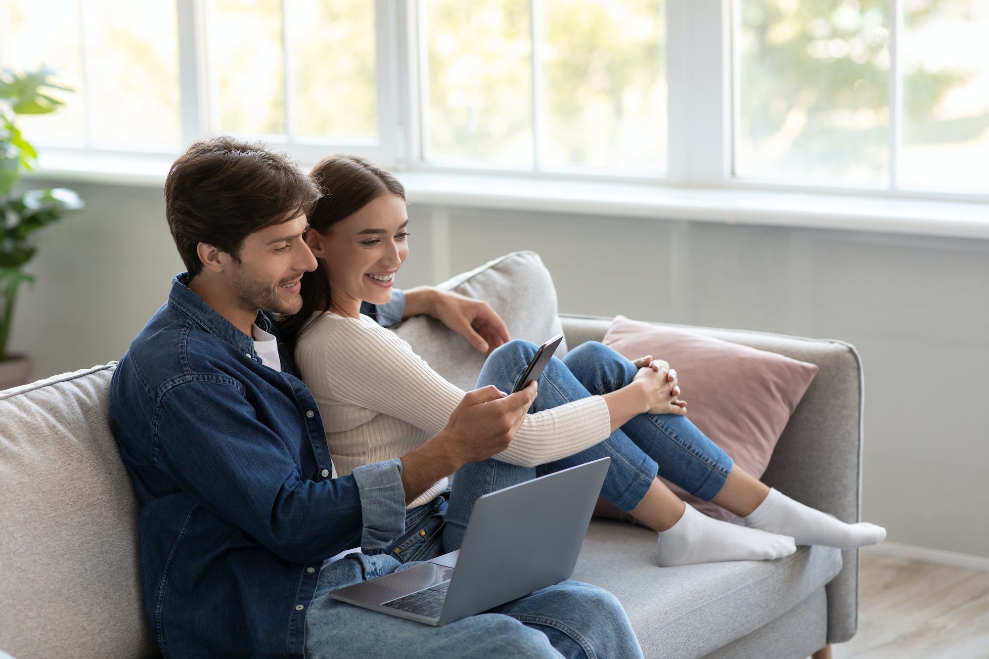 Happy millennial european male and female look at smartphone and call online in living room interior